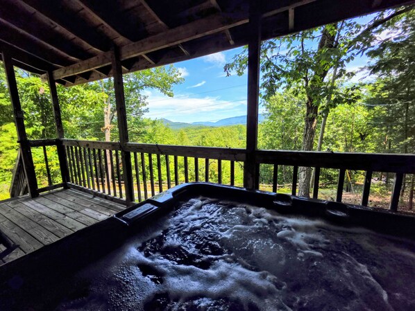 A hot tub with a wooden deck and a view of a forest and mountains.