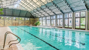 Una piscina interior con techos y ventanas de vidrio transparente, rodeada de sillones de descanso y plantas en macetas.