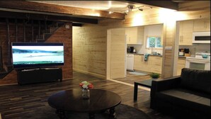 En moderne stue med et fladskærms-TV, et mørkt træsofabord med blomster og en sofa. Stuen har trægulv og et køkkenområde med hvide skabe og en komfur.