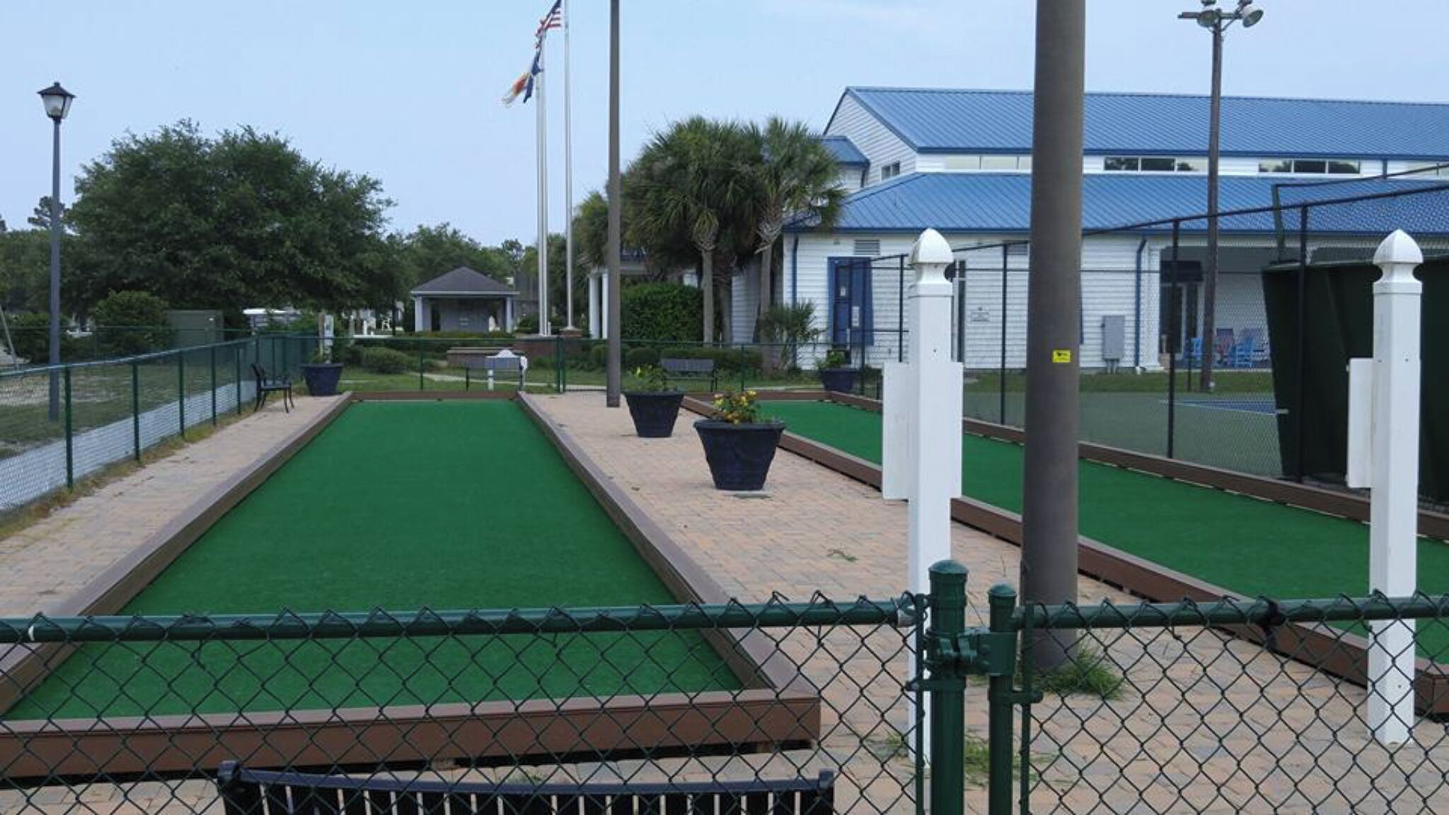 A miniature golf course with a green playing surface, surrounded by a chain-link fence, and a building with a blue roof in the background.