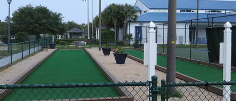 A miniature golf course with a green playing surface, surrounded by a chain-link fence, and a building with a blue roof in the background.
