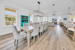 A spacious dining area with a long wooden table and grey upholstered chairs, a teal cabinet, and a ceiling fan.
