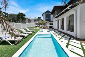 A swimming pool with lounge chairs, surrounded by a white fence and greenery.