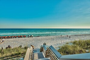 A beach with a wooden walkway leading to the shore, a row of umbrellas, and people walking along the beach.