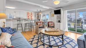 A spacious living area with a sofa, a round coffee table, and two wicker chairs. There is a ceiling fan, a TV mounted on the wall, and a large window offering a view of the outside. - Sunkissed 2: Comfortable Cottage Near Public Beach Access (Oak Island)