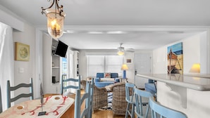 A modern kitchen with a dining area, featuring a large island, blue chairs, and a ceiling fan. - Sunkissed 2: Comfortable Cottage Near Public Beach Access (Oak Island)