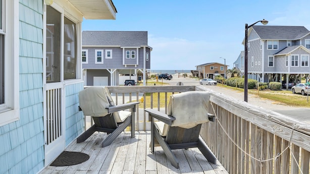 A wooden deck with two chairs, a railing, and a view of a residential street. - Sunkissed 2: Comfortable Cottage Near Public Beach Access (Oak Island)