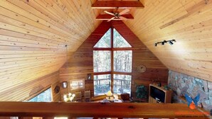 Een chalet met schuin dak, houten wanden, een groot raam, een plafondventilator, een klok, een open haard en een stenen muur.