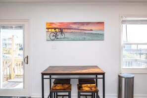 A room with a wooden table and benches, a painting of a bicycle on a beach, and a window with a view.