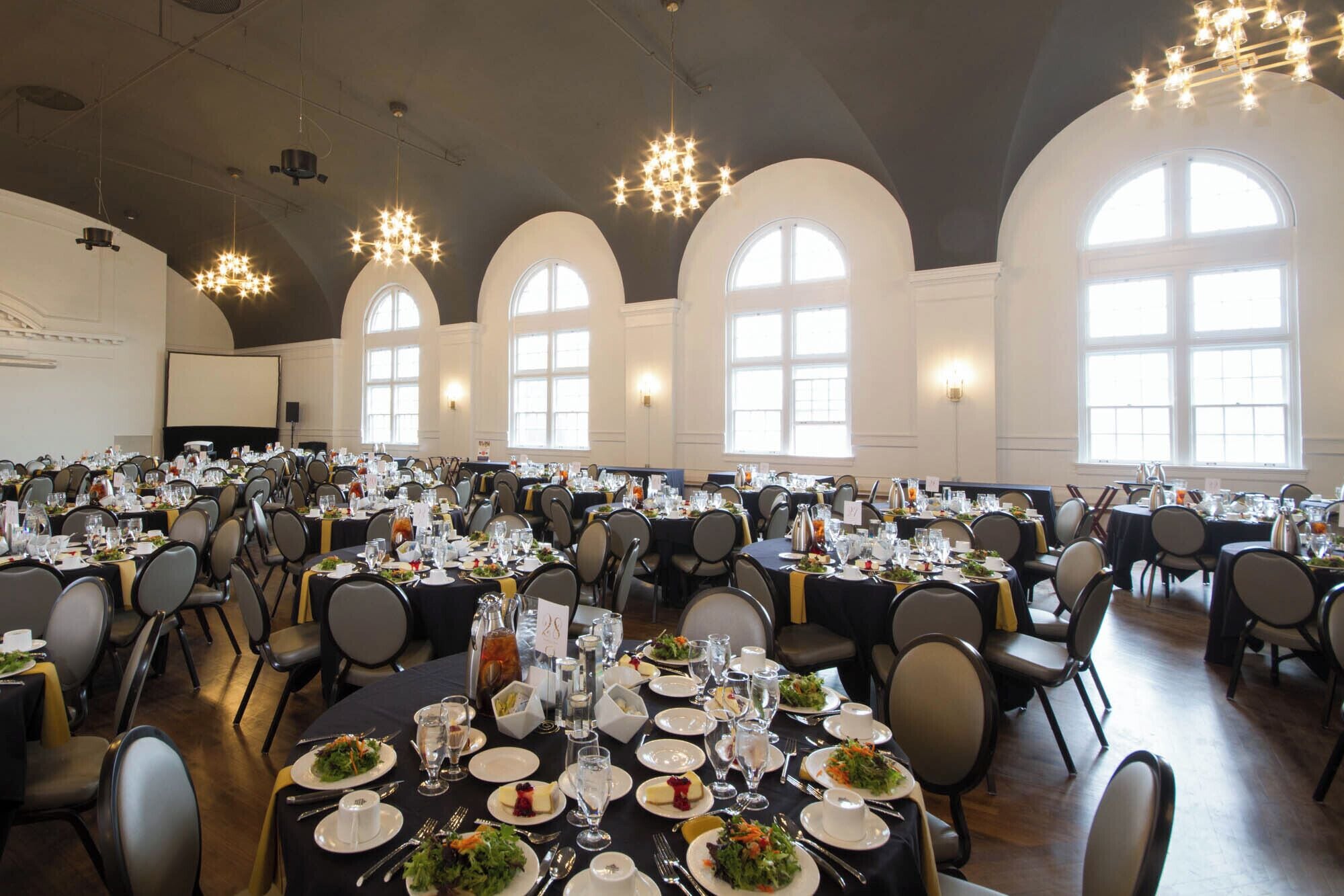 Een feestzaal met ronde tafels gedekt voor een diner, grote gebogen ramen en kroonluchters.