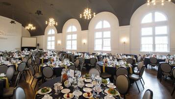 Een feestzaal met ronde tafels gedekt voor een diner, grote gebogen ramen en kroonluchters.