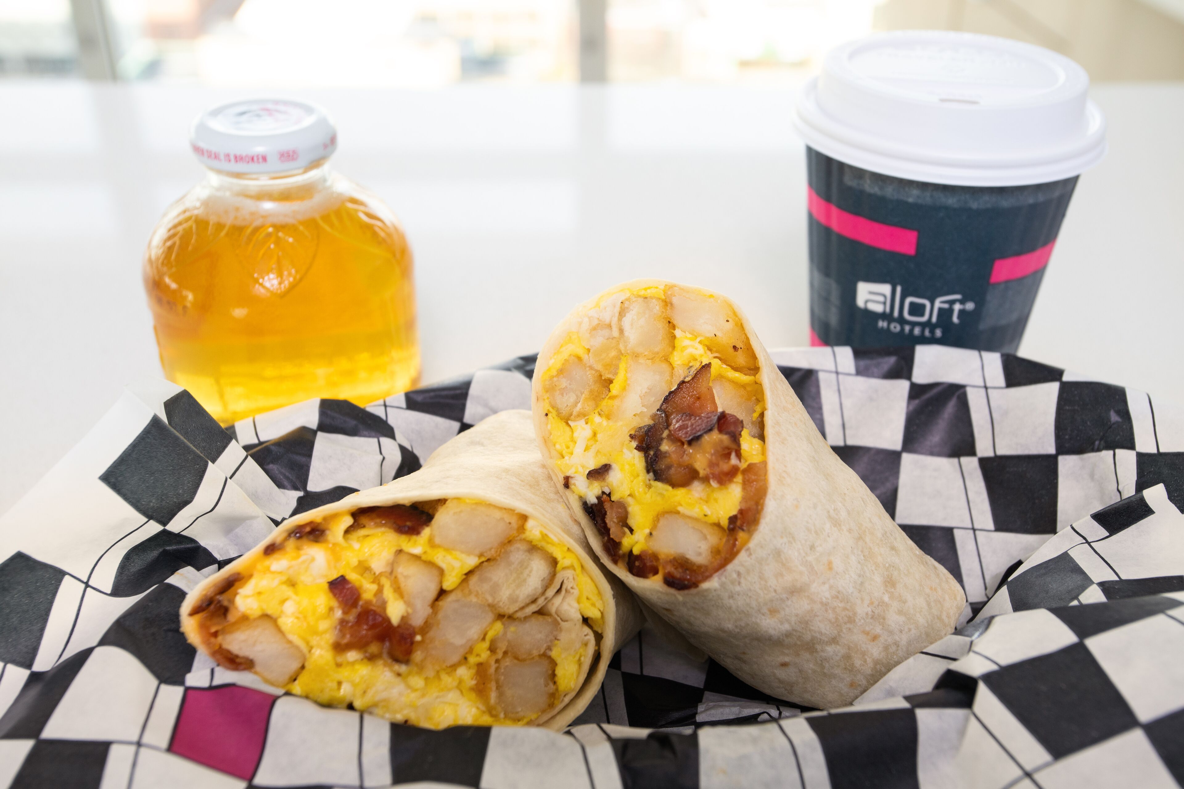 Un burrito dont on peut voir la garniture, une bouteille de miel et une tasse de café avec un couvercle.