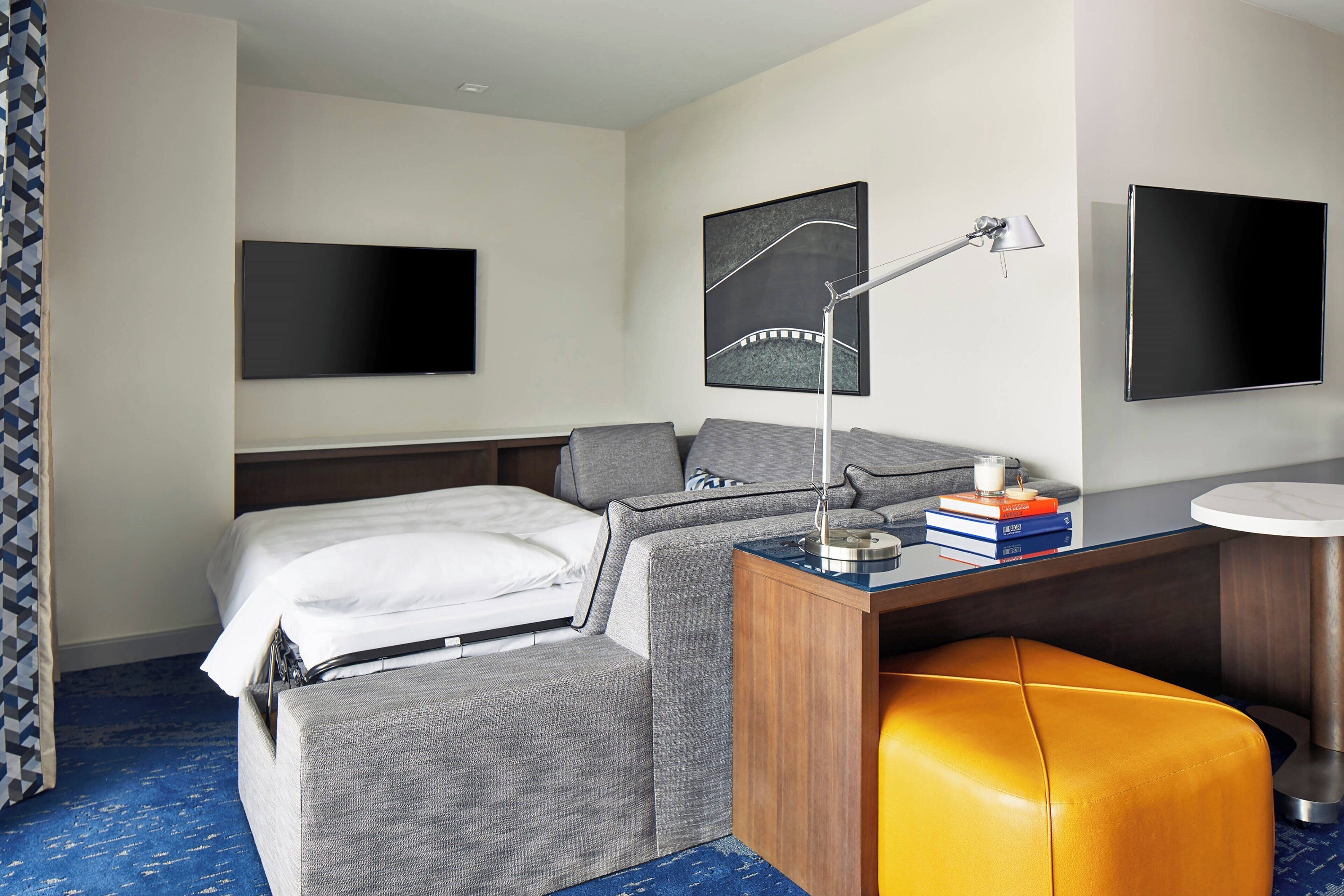 Quarto de hotel moderno com cama, escrivaninha, poltrona e televisão.