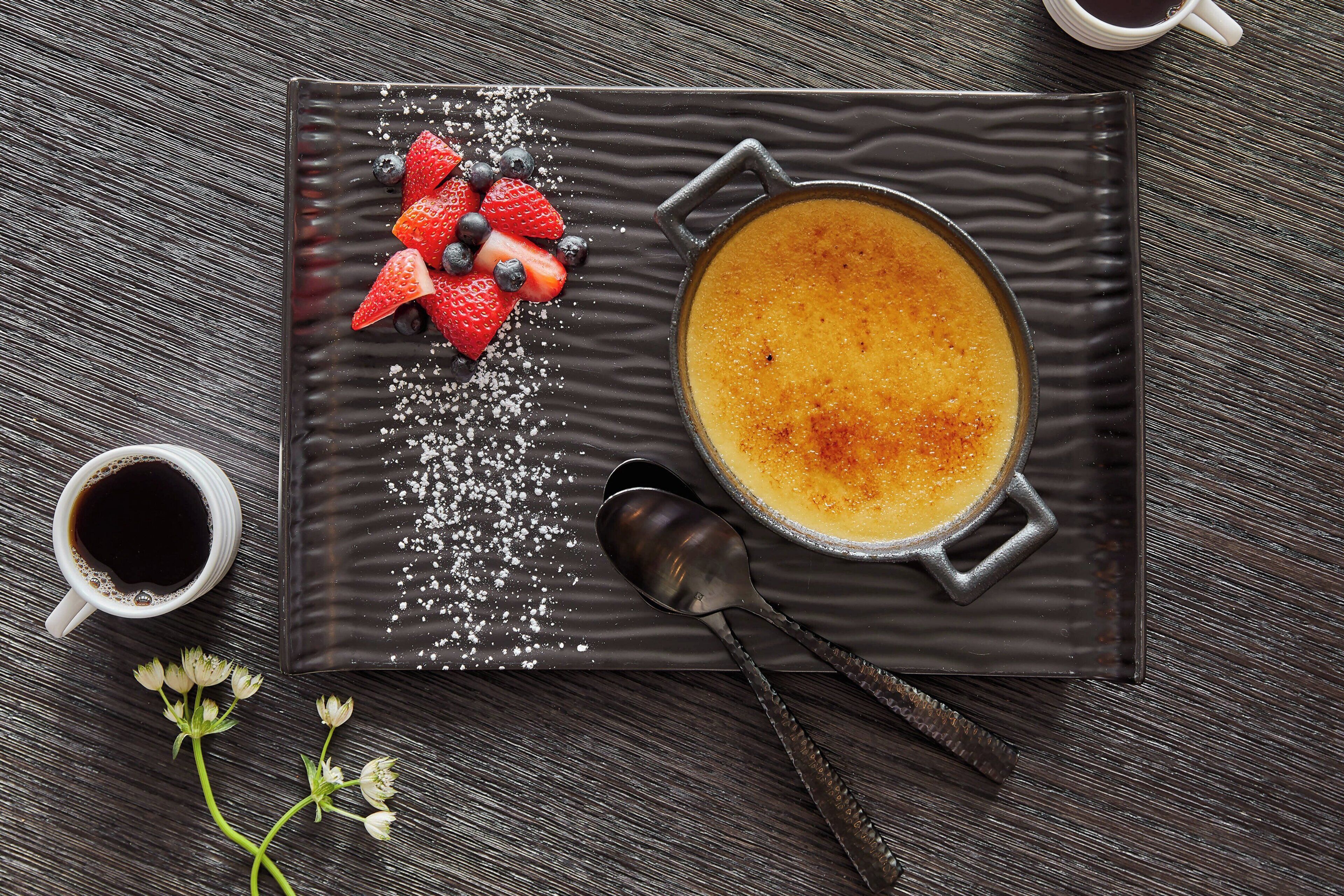 Une assiette de dessert contenant une crème brûlée, des fraises et des myrtilles, accompagnée d’une tasse de café et d’une cuillère, le tout posé sur une surface sombre et texturée.