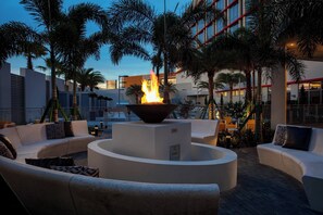 A modern outdoor lounge area with a central fire feature, surrounded by white sofas and palm trees. - The Daytona, Autograph Collection (Daytona Beach)