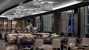 Un ristorante moderno con soffitti alti, lampade a sospensione a righe e una motocicletta esposta al centro. I tavoli sono apparecchiati con bicchieri e posate, e l'ambiente presenta un'ampia finestra con tende.