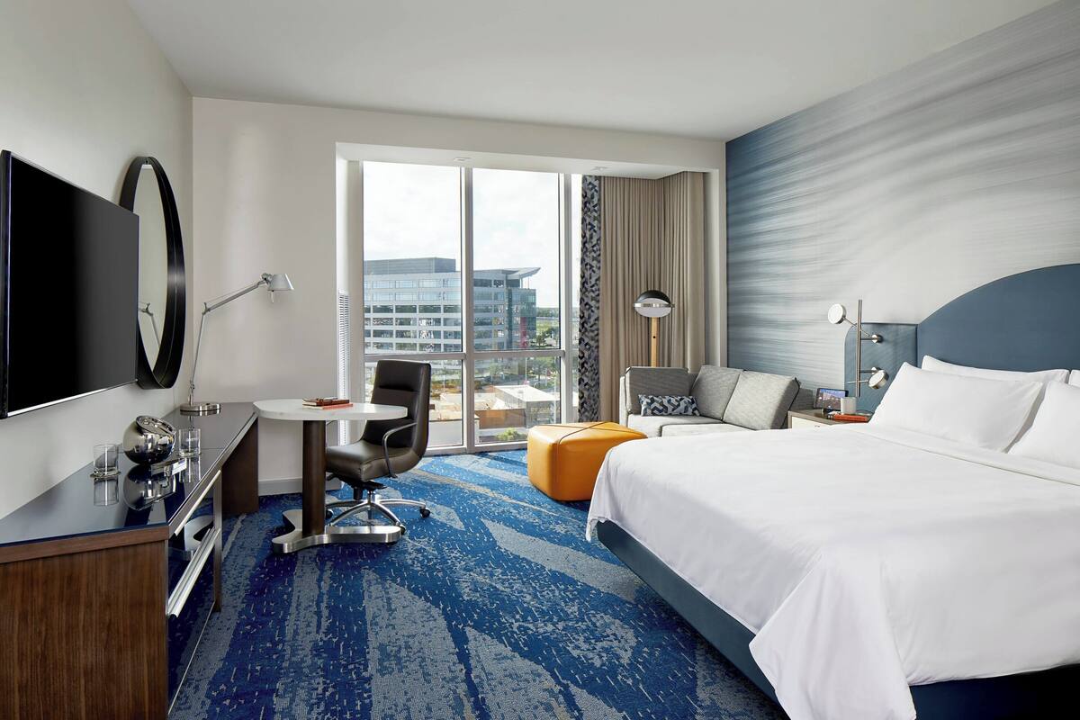 A hotel room with a large bed, a desk with a chair, a TV, and a view of the city through the window.