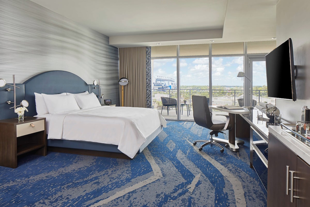 A hotel room with a large bed, a desk with a chair, a TV, and a view of a cityscape through large windows.