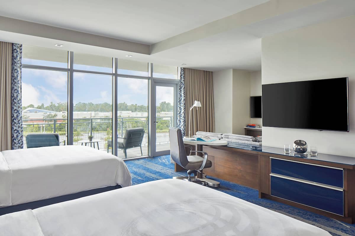 A hotel room with two beds, a desk with a computer, a TV, and a large window with a view of the city.