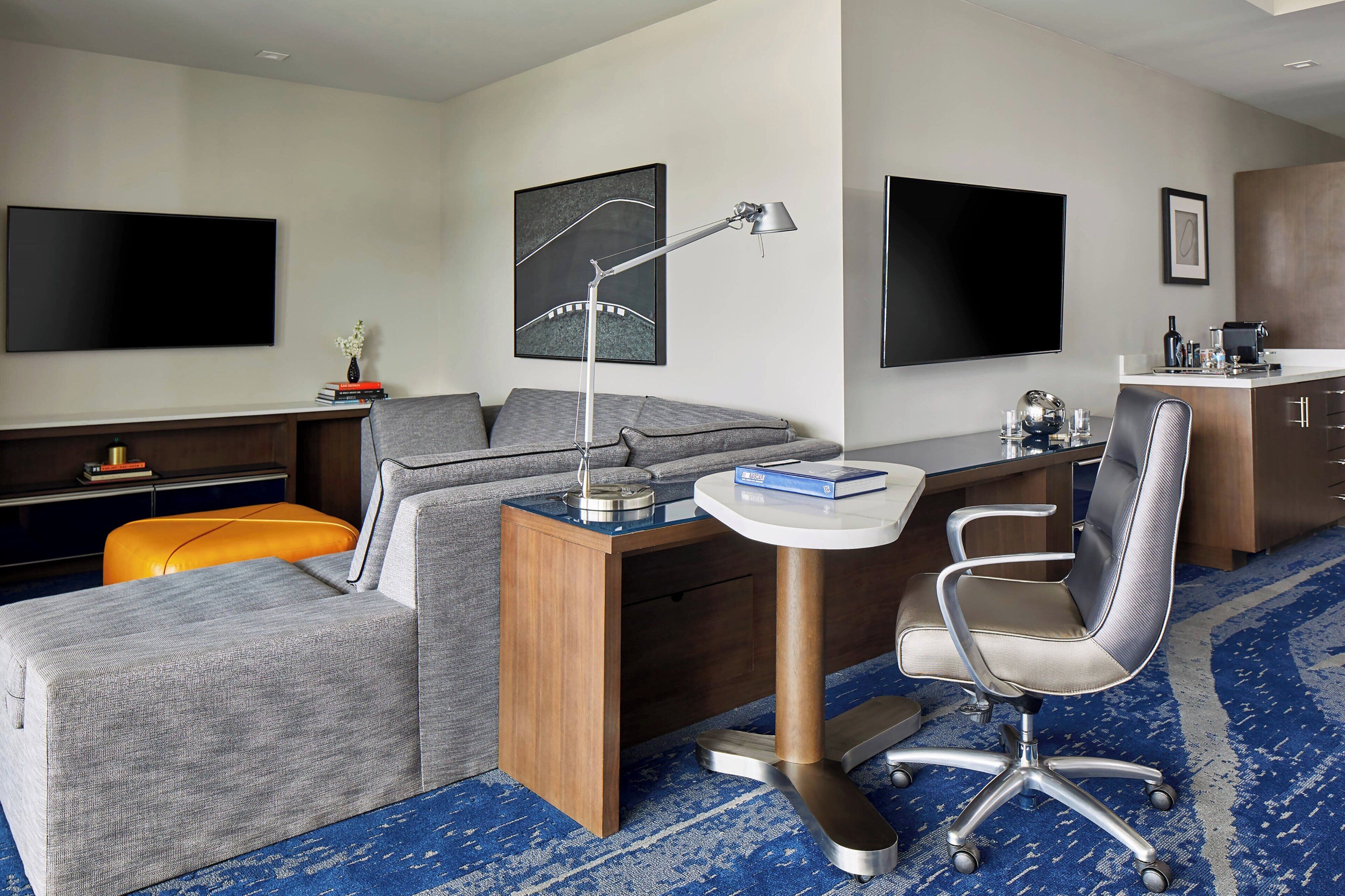 Una habitación de hotel moderna con sofá, escritorio, silla, televisor de pantalla plana, cafetera y un adorno mural.