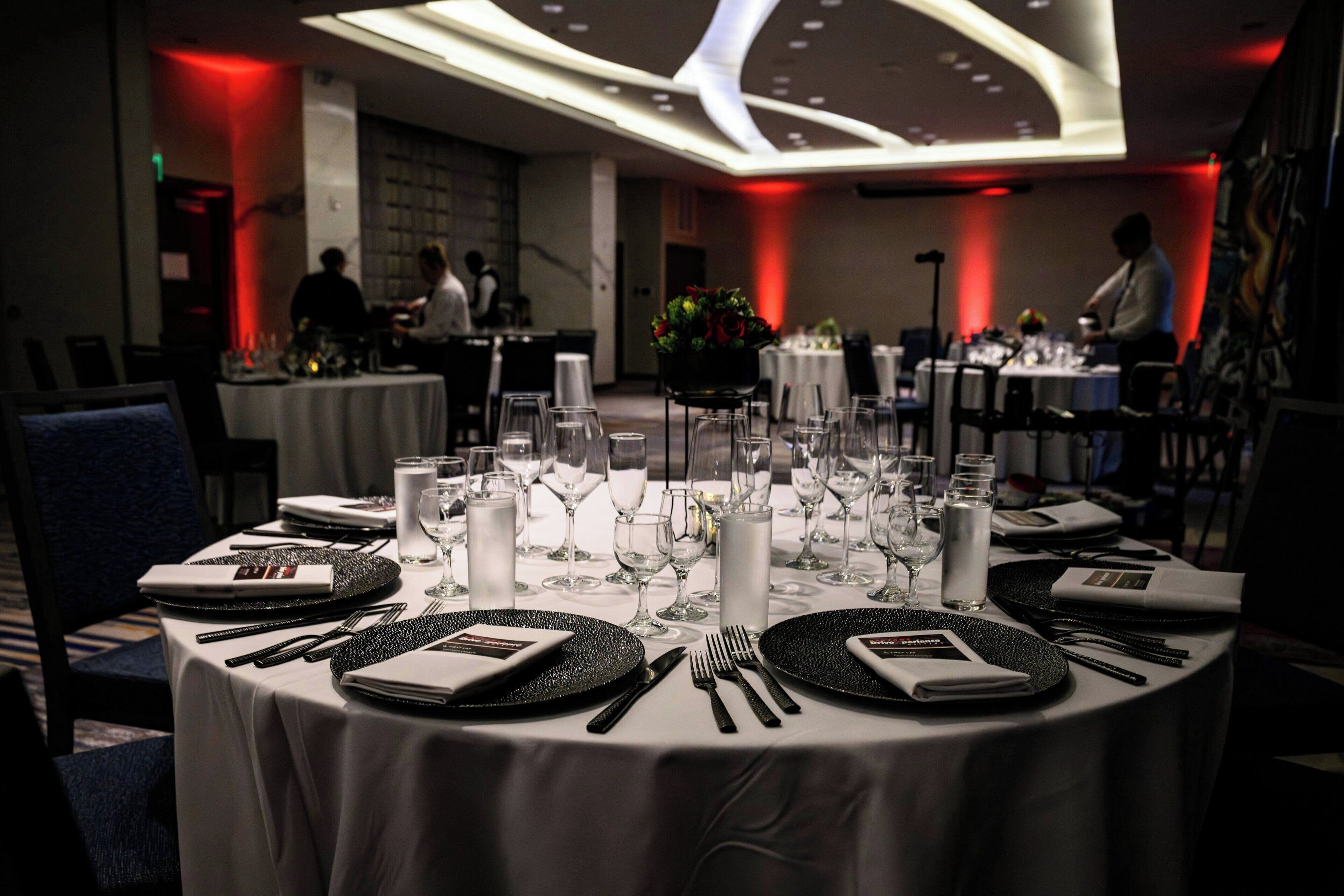 A dining area with round tables set for a formal meal, featuring white tablecloths, black plates, and silverware.