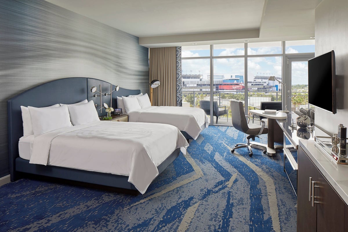 A hotel room with two beds, a desk, a chair, a TV, and large windows offering a view of the city.