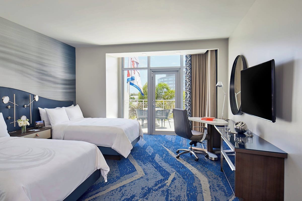 A hotel room with two beds, a desk, a chair, a TV, and a balcony with a view of the outside.
