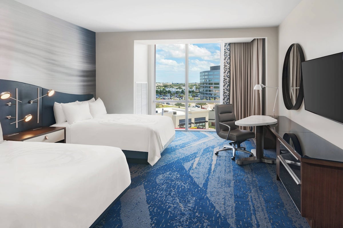 A hotel room with two beds, a desk, a chair, a TV, and a large window with a city view.