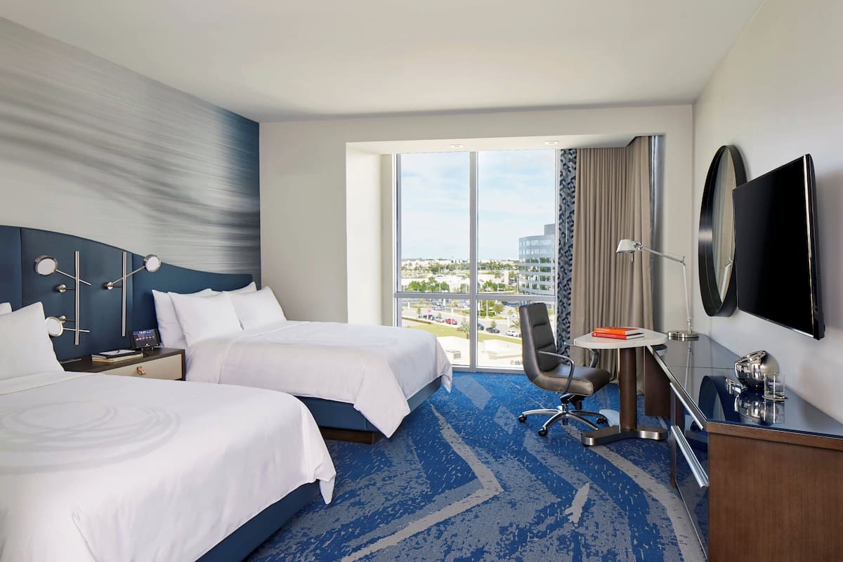 A hotel room with two beds, a desk, a chair, a TV, and a large window offering a city view.