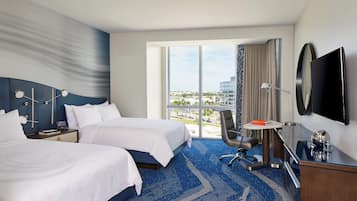 A hotel room with two beds, a desk, a chair, a TV, and a large window offering a city view.