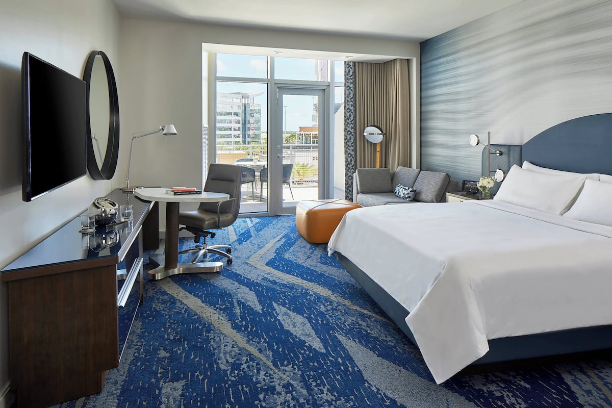 A hotel room with a large bed, a desk with a flat-screen TV, a chair, a small table, and a view of the city through the balcony doors.