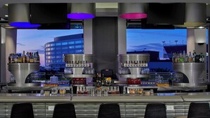 Un'area bar dal design moderno, con un grande schermo che mostra un panorama urbano, diversi banconi bar con bottiglie e bicchieri vari, e quattro sgabelli da bar neri.