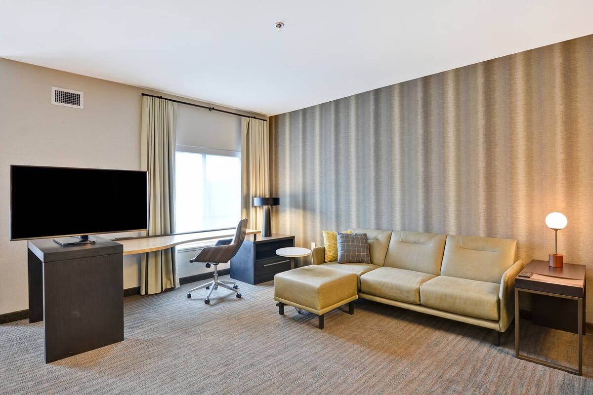 Une chambre d’hôtel équipée d’un grand téléviseur à écran plat, d’un canapé, d’un pouf, d’un bureau avec une chaise et d’une lampe.