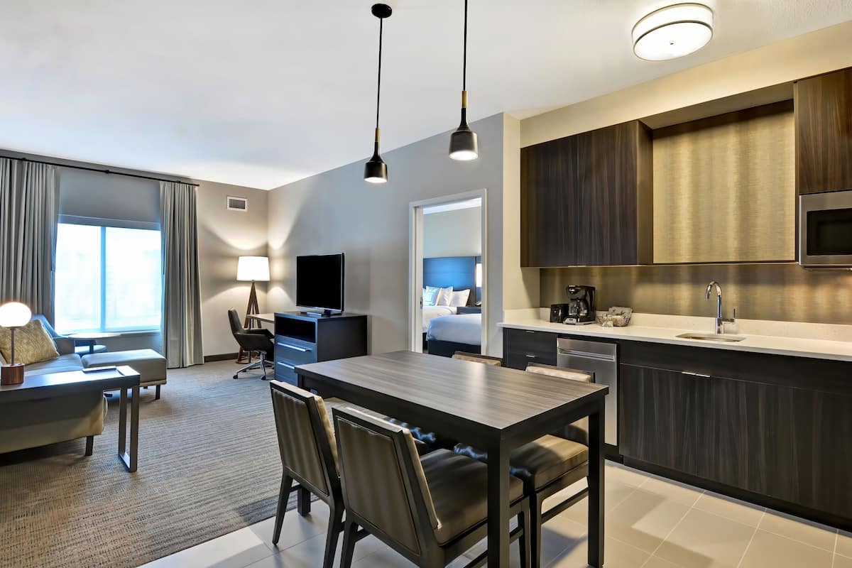 Une chambre d’hôtel moderne dotée d’un coin repas, d’une kitchenette et d’une grande fenêtre avec des rideaux.