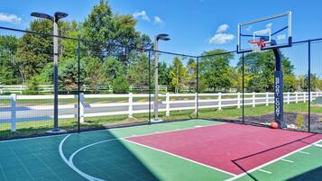 An outdoor basketball court with a green and red playing surface, surrounded by a white fence and trees.