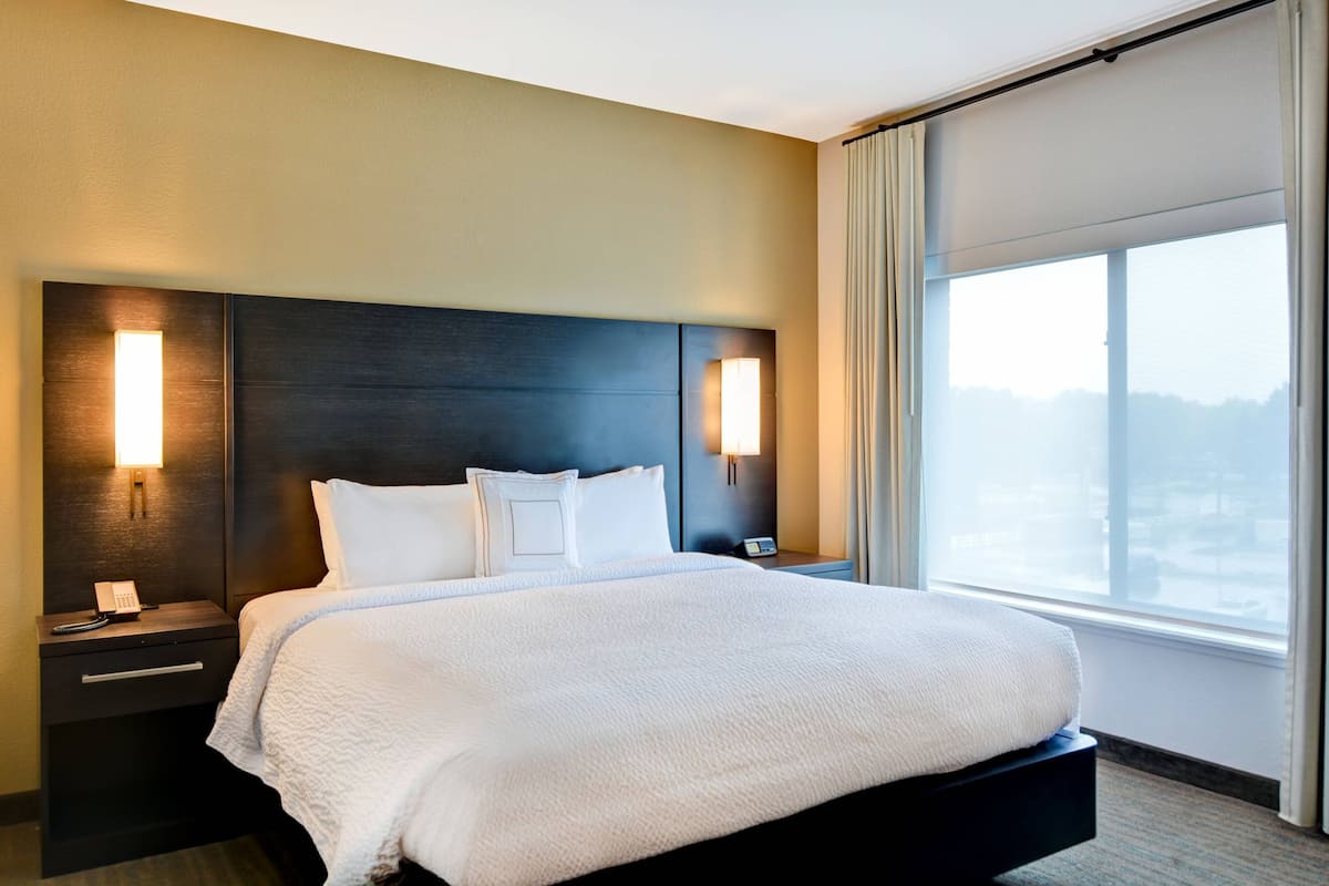 Une chambre d’hôtel avec un grand lit, deux tables de chevet, une petite table et une fenêtre donnant sur un beau paysage.