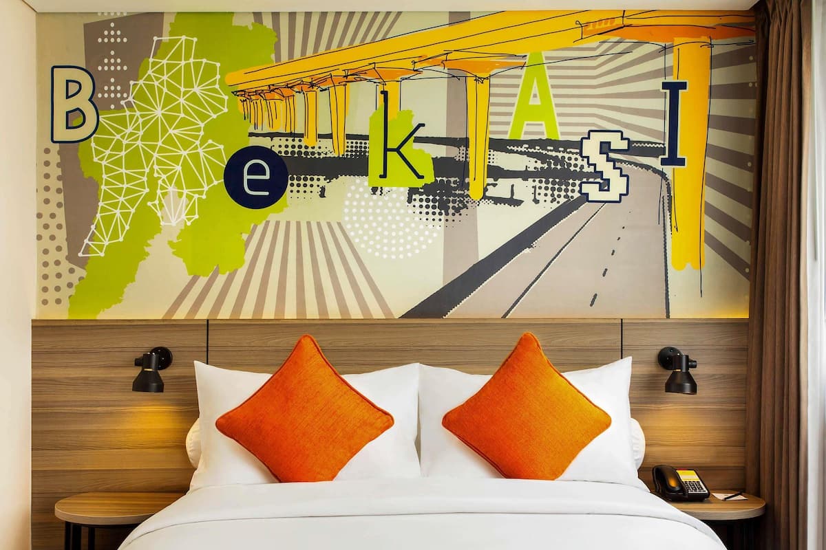 A hotel room with a bed, two orange pillows, a wooden headboard, a wall art piece, and a bedside table with a phone.