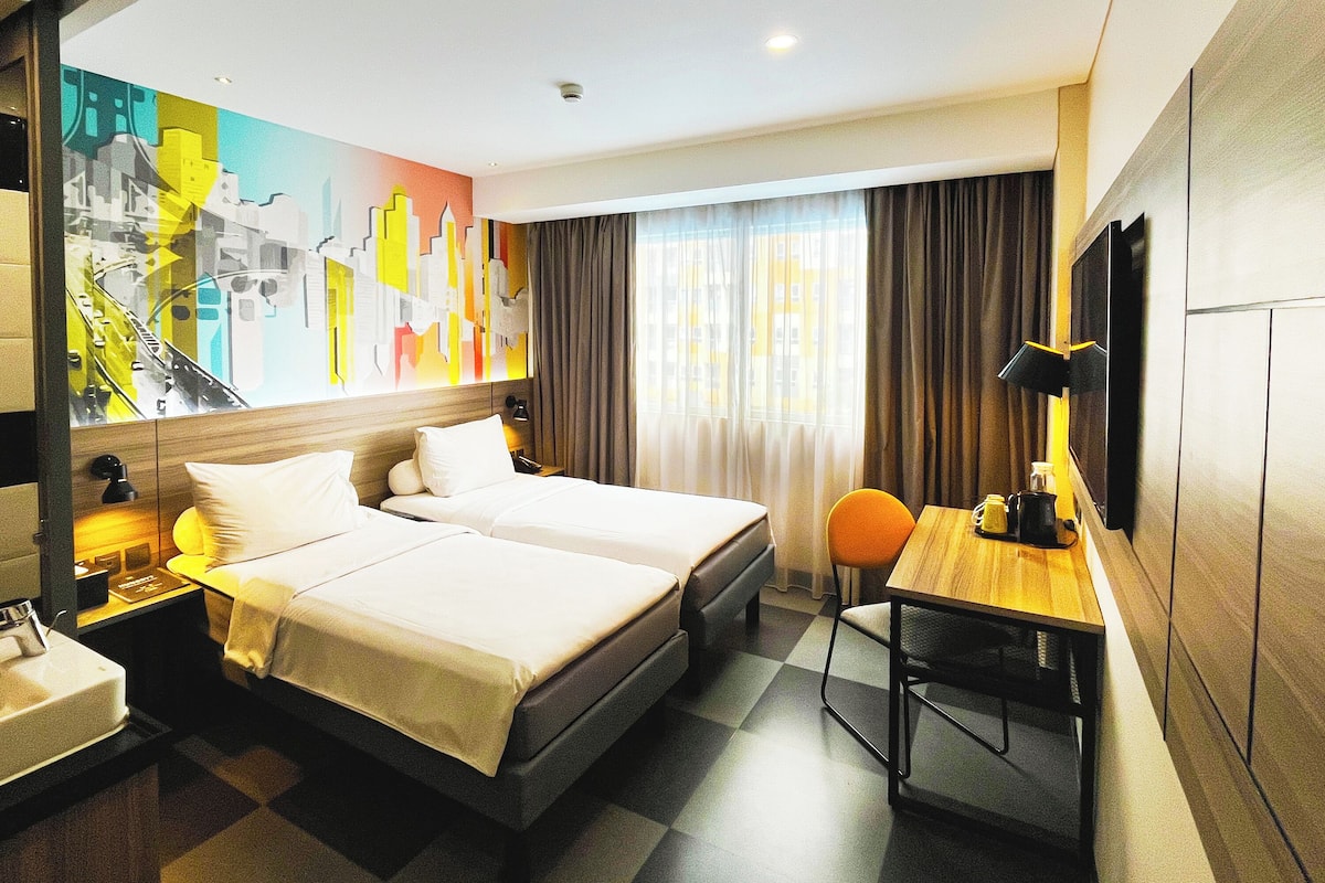 A hotel room with two beds, a desk, a chair, a desk lamp, and a large window with curtains.