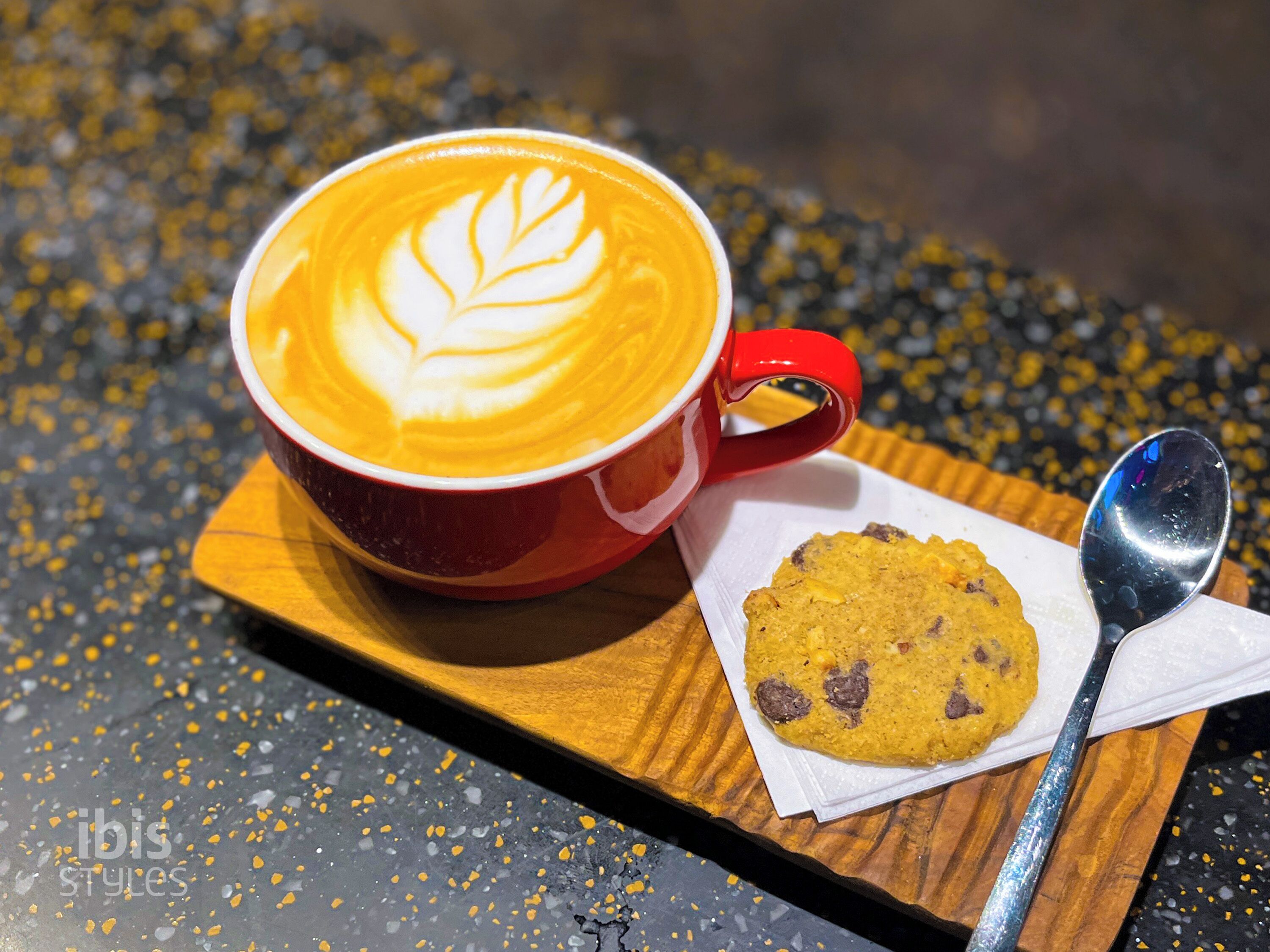 A red cup of coffee with latte art on a wooden tray, accompanied by a cookie and a spoon.
