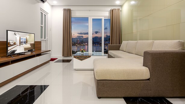 A modern living room with a sofa, a flat-screen TV, a glass coffee table, and a view of a cityscape through large windows.