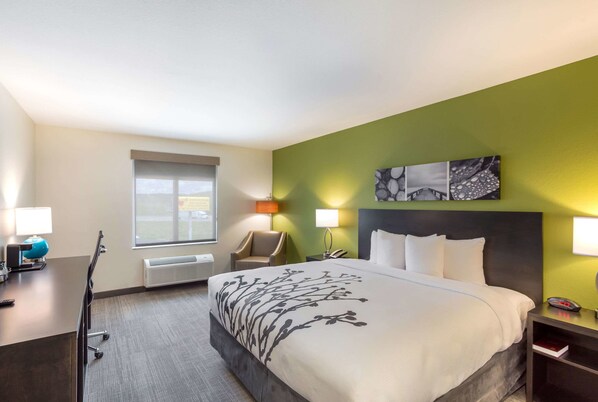 A hotel room with a large bed, a desk with a lamp, a chair, a window, and a wall-mounted artwork.