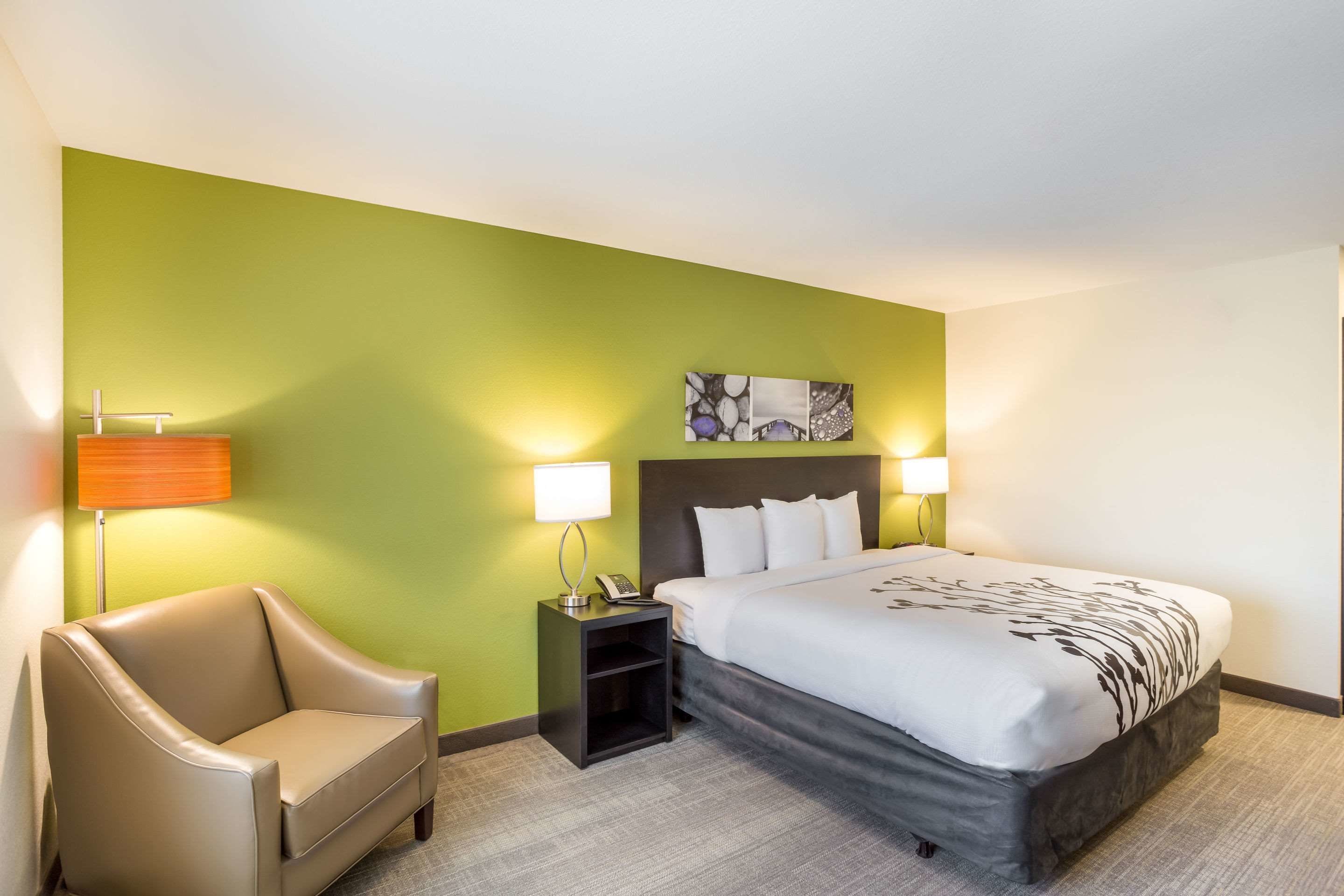Hotellihuone, jossa on sänky, tuoli, yöpöytä ja seinään kiinnitetty valaisin.