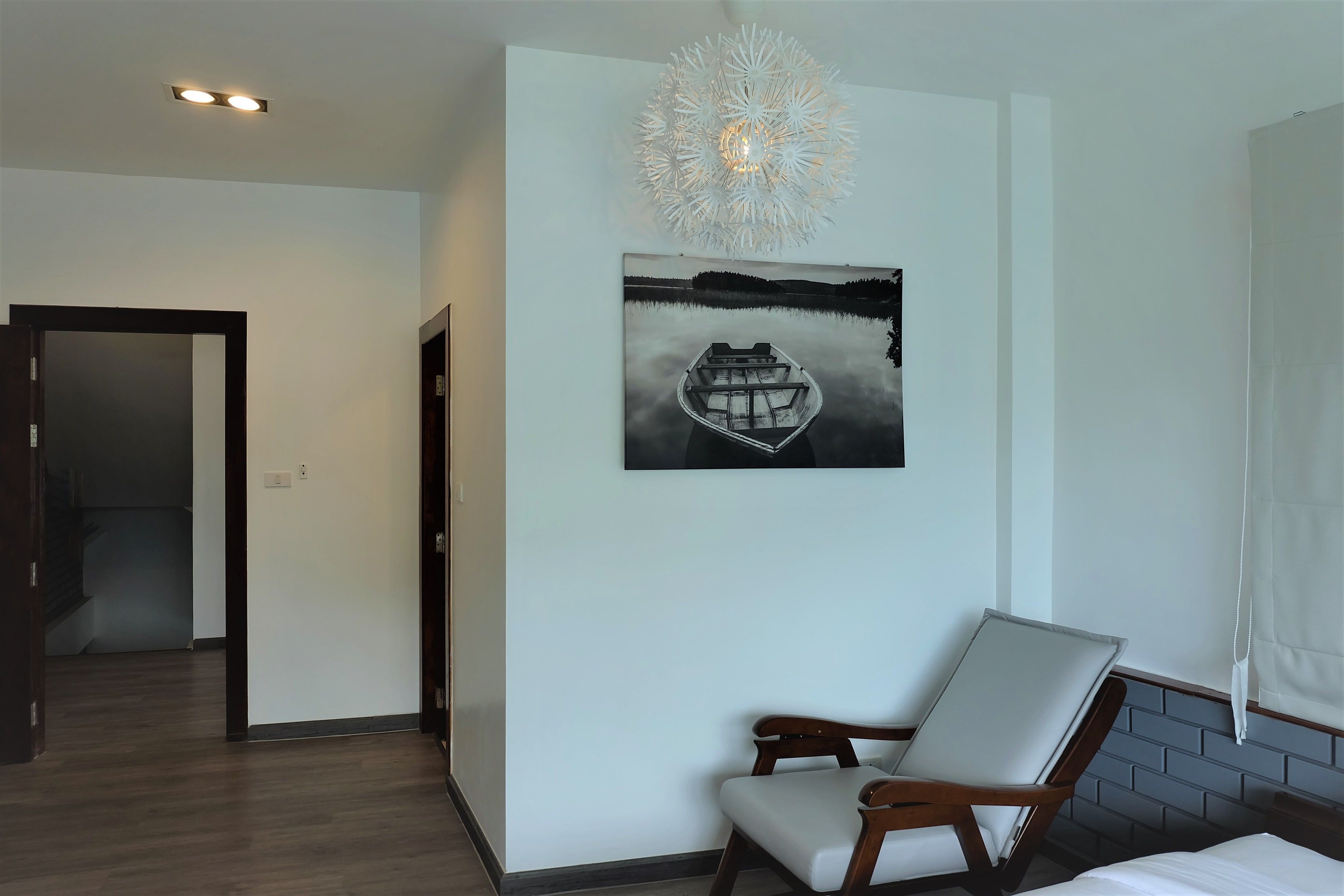 A hotel room with a wooden floor, a bed with white linens, a wooden chair with a white cushion, a wall-mounted picture of a boat on water, and a ceiling light fixture.