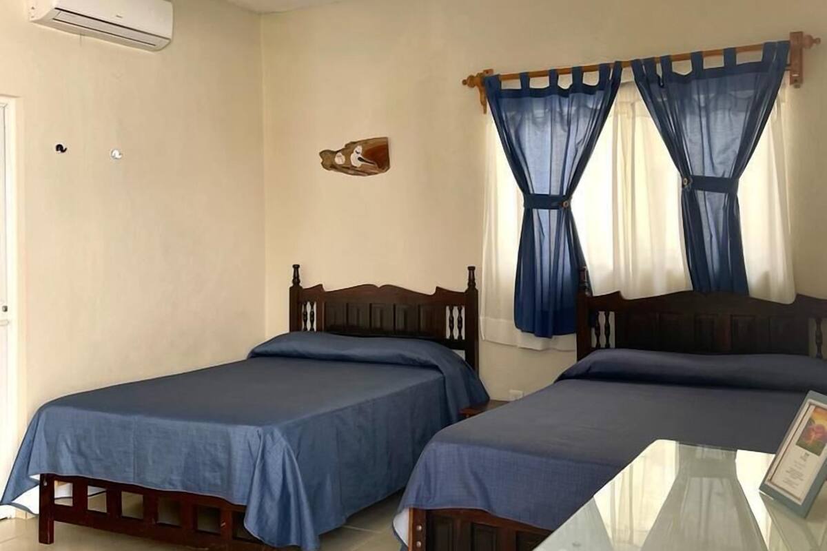 Una habitación con dos camas de madera, un ventilador de techo, un equipo de aire acondicionado y una mesa de vidrio.