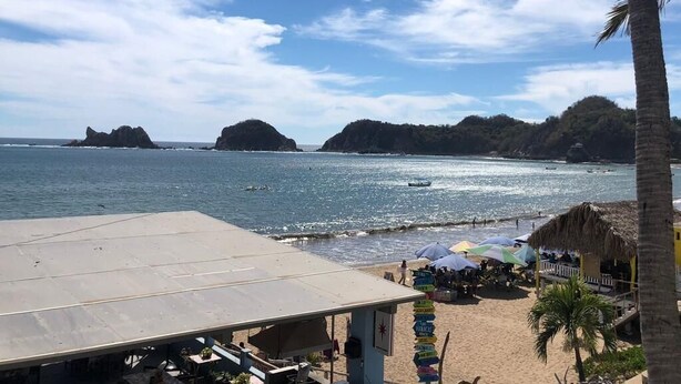 A beach with a thatched-roof structure, palm trees, and a clear view of the sea and distant islands. - Bungalows Las Hamacas (San Patricio Melaque)