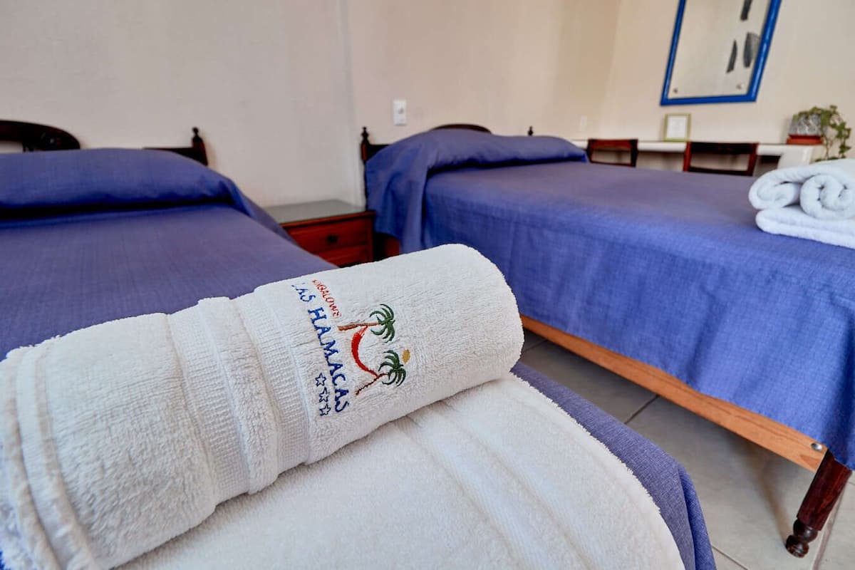 Habitación de hotel con dos camas, cada una con colcha y almohadas azules. Una toalla blanca con un logotipo está sobre una silla.