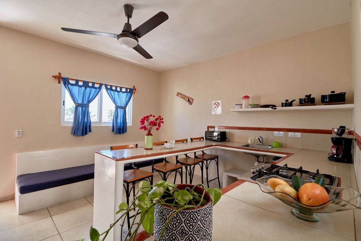 Cocina compacta con una isla central, ventilador de techo, ventana con cortinas azules y una banca.