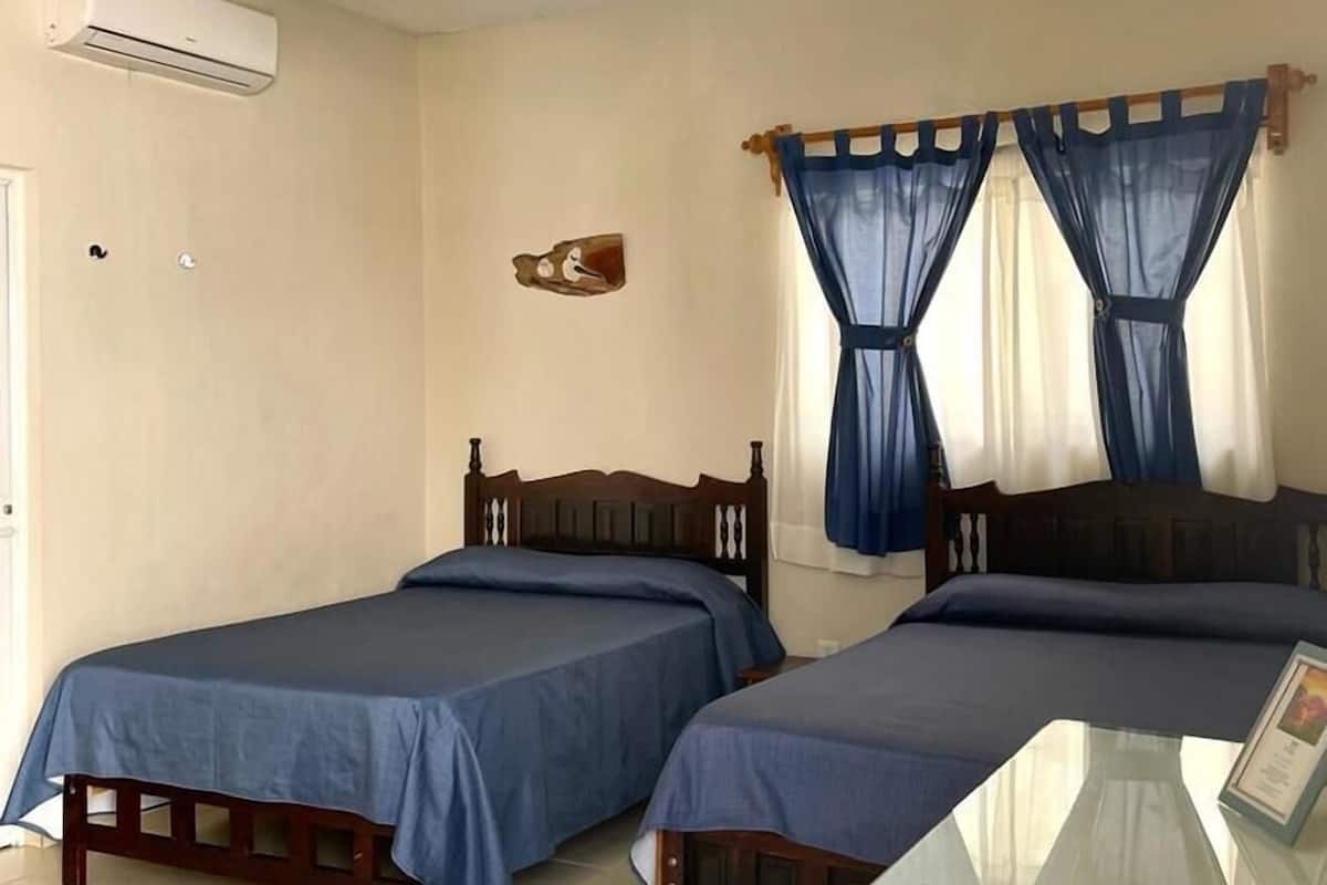Una habitación con dos camas individuales, ventilador de techo, equipo de aire acondicionado, una mesita pequeña y una ventana con cortinas azules.