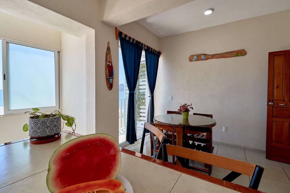 Un comedor con una mesa redonda, sillas, una planta en maceta y una sandía grande sobre la mesa.