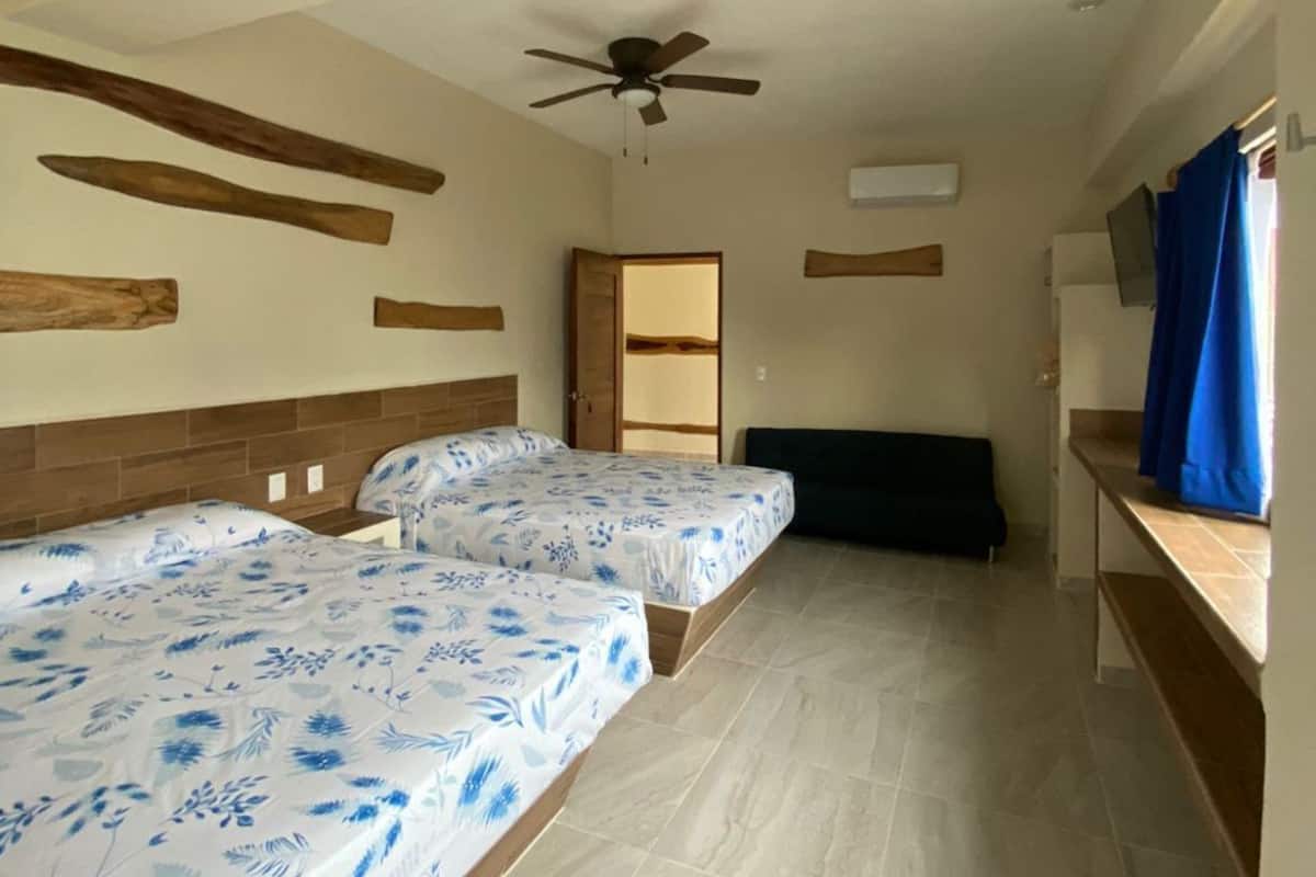 Una habitación con dos camas, ventilador de techo, cabecera de madera, un banco, televisión y ventana con cortinas.